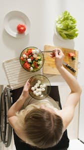 Top view of a woman in a wheelchair preparing a healthy salad with fresh ingredients in a kitchen.